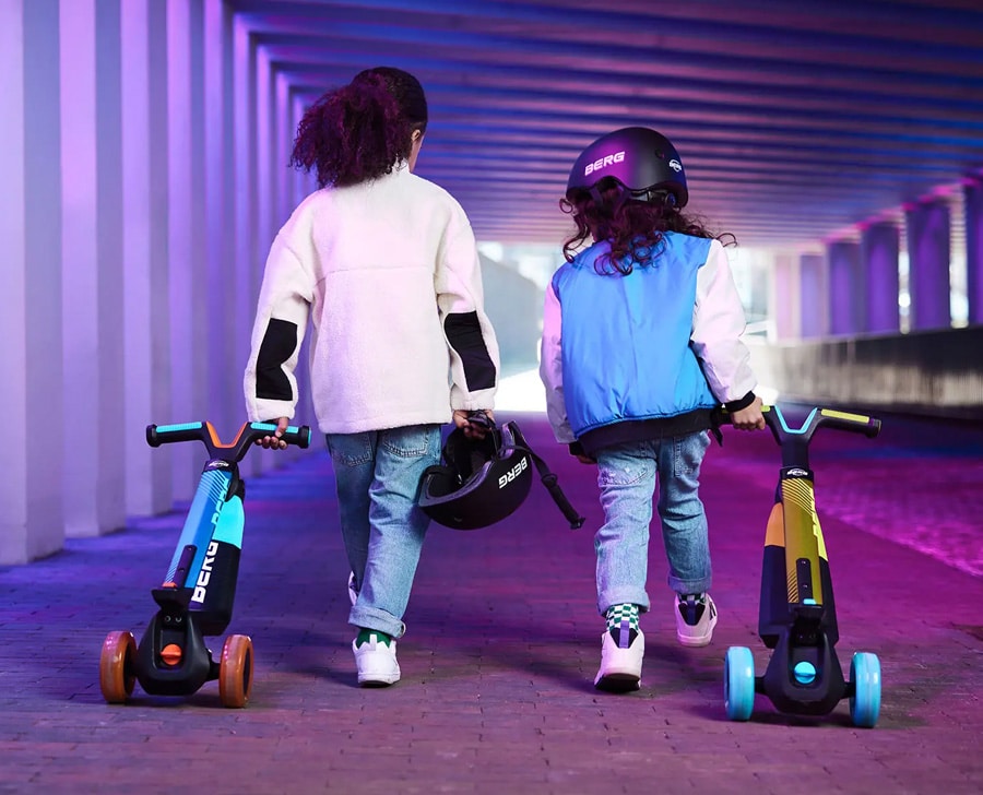 Two Girls Holding A Blue And A Yellow BERG Nexo Scooters With Light-Up Wheels